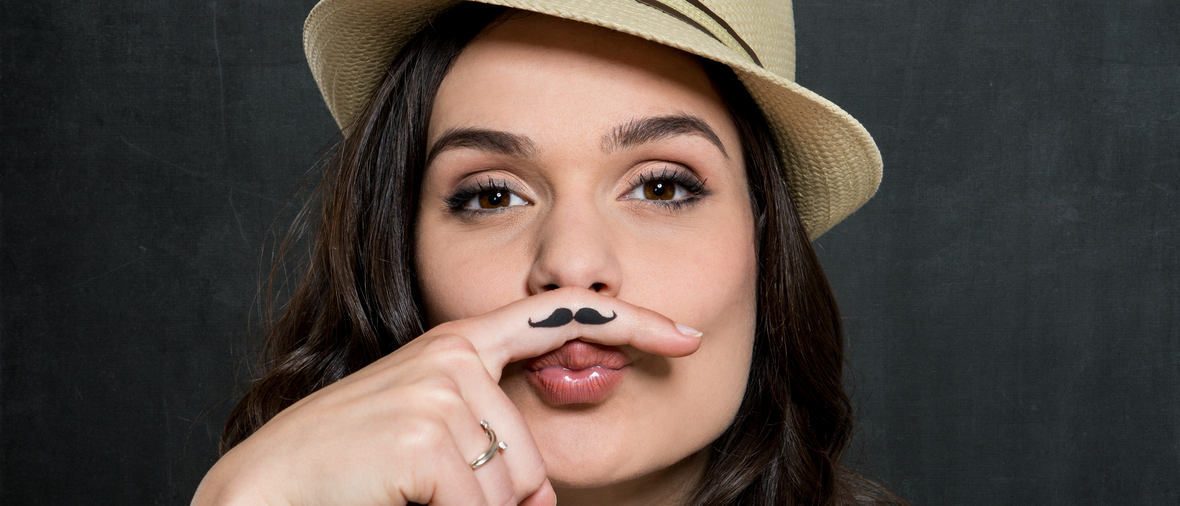 Portrait Of Young Woman In Hat With Painted Moustache Over Gray Background
